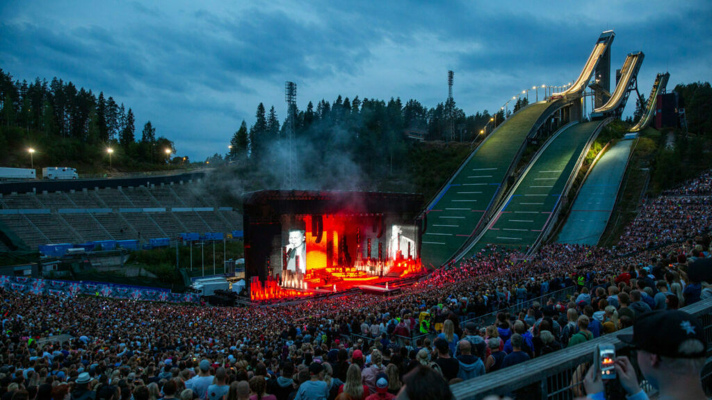 Yleisömassa suuren tapahtulavan edessä, taustalla kolme mäkihypyn hyppyrimäkeä.