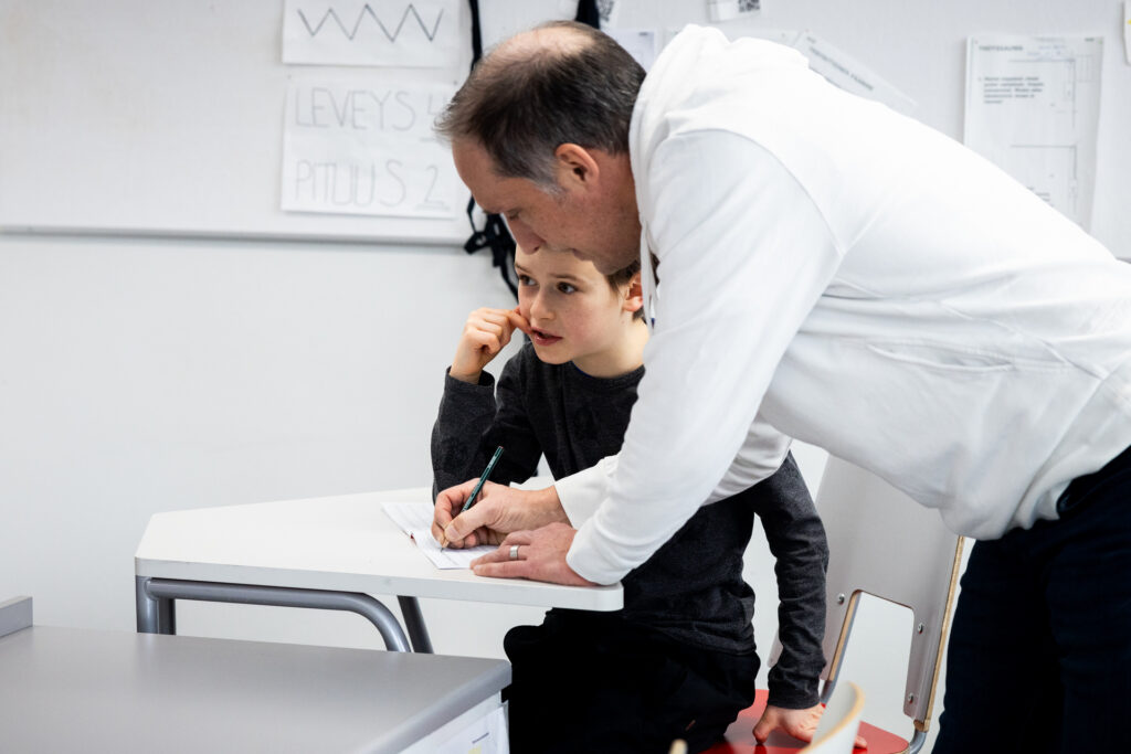 Esa Liukkonen on kumartunut pulpetin ylle kirjoittamaan jotain oppilaan paperille. Pulpetin ääressä on 4-luokkalainen oppilas.