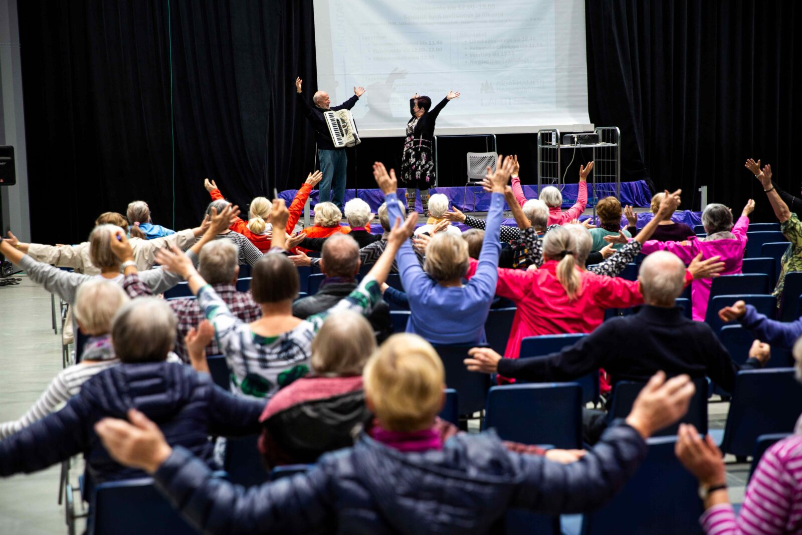 Senioreiden hyvinvointipäivässä yleisö toistaa lavalta näytetyn liikkeen, jossa avataan kädet levälleen.